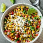 A white bowl filled with Mexican Street Corn Salad, topped with cheese, diced red peppers, onions, herbs, and a lime wedge, sits on a grey surface next to lime slices and a spoon.