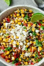 A white bowl filled with Mexican Street Corn Salad, topped with cheese, diced red peppers, onions, herbs, and a lime wedge, sits on a grey surface next to lime slices and a spoon.
