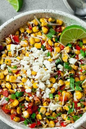 A white bowl filled with Mexican Street Corn Salad, topped with cheese, diced red peppers, onions, herbs, and a lime wedge, sits on a grey surface next to lime slices and a spoon.