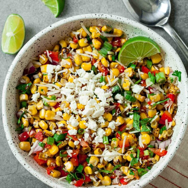 A white bowl filled with Mexican Street Corn Salad, topped with cheese, diced red peppers, onions, herbs, and a lime wedge, sits on a grey surface next to lime slices and a spoon.