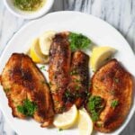 Three seasoned and cooked Pan Seared Tilapia fillets are served on a white plate with lemon wedges and parsley garnish, perfect for a delicious fish dinner.