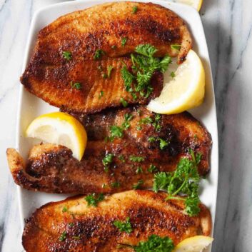 Three pieces of pan seared tilapia on a white plate, garnished with parsley and lemon wedges, served with a small bowl of buttery lemon sauce and extra lemon slices.
