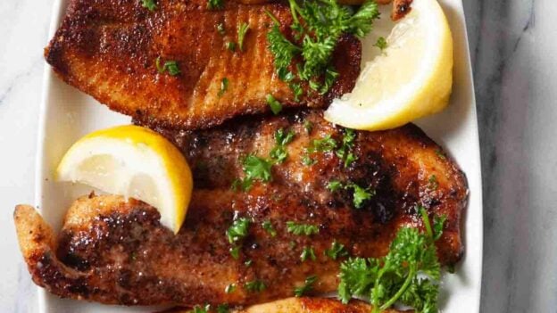 Three pieces of pan seared tilapia on a white plate, garnished with parsley and lemon wedges, served with a small bowl of buttery lemon sauce and extra lemon slices.