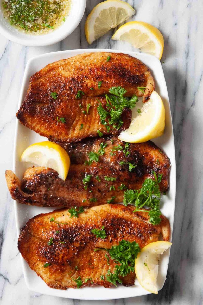 Three pieces of pan seared tilapia on a white plate, garnished with parsley and lemon wedges, served with a small bowl of buttery lemon sauce and extra lemon slices.