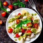 A plate of Tortellini Pasta Salad topped with cherry tomatoes, mozzarella balls, pesto sauce, and fresh basil sits next to a fork and basil leaves on a dark surface.