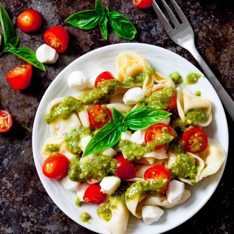 A plate of Tortellini Pasta Salad topped with cherry tomatoes, mozzarella balls, pesto sauce, and fresh basil sits next to a fork and basil leaves on a dark surface.