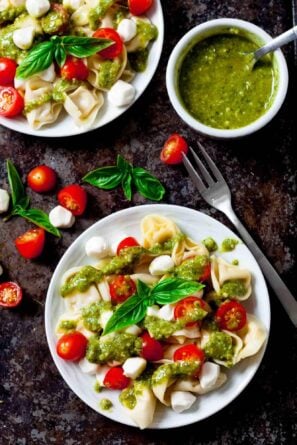 Plates of Tortellini Pasta Salad with cherry tomatoes, Mozzarella balls, fresh basil, and vibrant pesto sauce, served with a fork and an extra bowl of pesto.