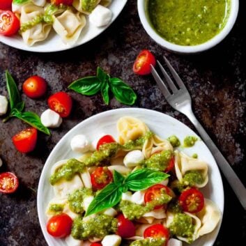 Plates of Tortellini Pasta Salad with cherry tomatoes, Mozzarella balls, fresh basil, and vibrant pesto sauce, served with a fork and an extra bowl of pesto.