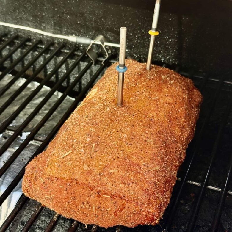 Pork loin with dry rub being smoked. Meat thermometer inserted in it to check the temperature.
