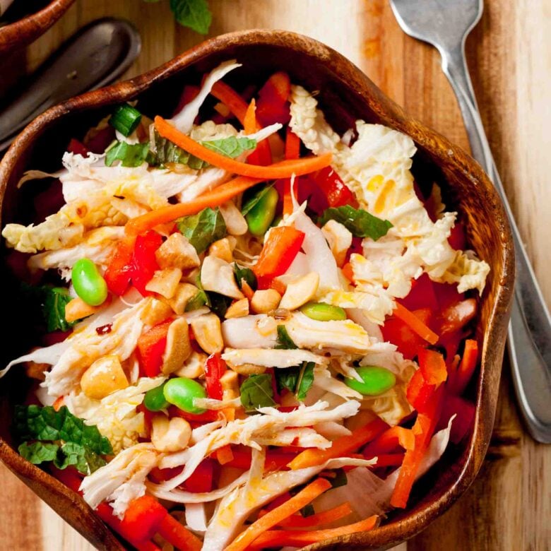 A wooden bowl filled with Vietnamese Chicken Salad—shredded chicken, cabbage, carrots, edamame, red bell pepper, chopped peanuts, and fresh herbs—all tossed with a tangy dressing. A fork rests beside this healthy salad.