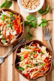 Two wooden bowls filled with Vietnamese Chicken Salad—a healthy salad of shredded chicken, carrots, bell peppers, edamame, cabbage, chopped peanuts, and herbs—sit on a wooden surface, with a fork and mint leaves nearby.