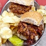 Pulled pork sandwiches on whole wheat buns served with potato chips and pickle slices on a metal tray.