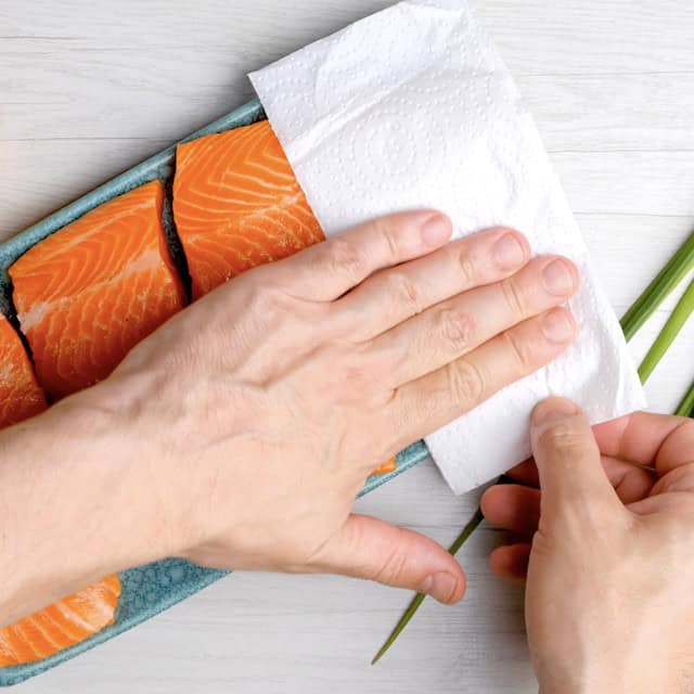 A person uses a paper towel to pat dry raw salmon fillets arranged in a blue dish on a light-colored surface, preparing them for an easy grilled salmon dinner.