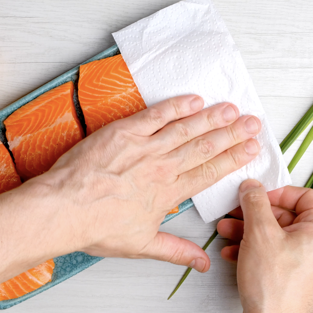 A person uses a paper towel to pat dry raw salmon fillets arranged in a blue dish on a light-colored surface, preparing them for an easy grilled salmon dinner.