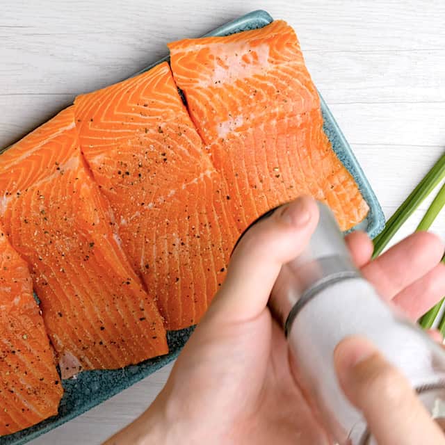 A hand sprinkles salt onto raw salmon fillets arranged on a blue rectangular plate, preparing for an Easy Grilled Salmon dinner.