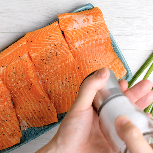 A hand sprinkles salt onto raw salmon fillets arranged on a blue rectangular plate, preparing for an Easy Grilled Salmon dinner.