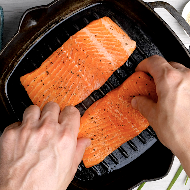 Two hands place two raw, seasoned salmon fillets onto a black grill pan, to make Easy Grilled Salmon recipe.
