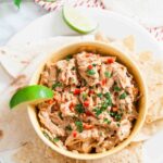 A bowl of creamy Mexican Chicken Salad dip topped with chopped herbs and red peppers, served with tortilla chips, flatbread, and a lime wedge on a white plate.