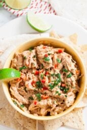 A bowl of Mexican Chicken Salad dip garnished with chopped herbs and red peppers, served with tortilla chips and a lime wedge.