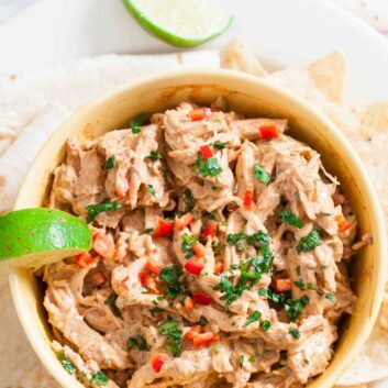 A bowl of Mexican Chicken Salad dip garnished with chopped herbs and red peppers, served with tortilla chips and a lime wedge.