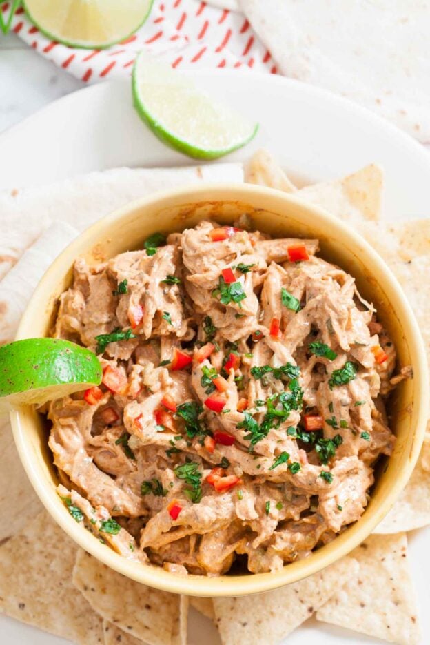 A bowl of Mexican Chicken Salad dip garnished with chopped herbs and red peppers, served with tortilla chips and a lime wedge.