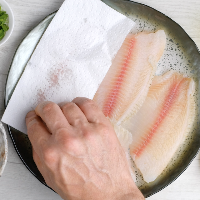 Tilapia fillet being patted dry with paper towels.