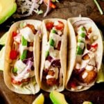 Three Baja Fish Tacos in soft tortillas, topped with crispy fish, sliced avocado, red onion, tomato, shredded lettuce, and a creamy sauce, arranged on a rustic surface with lime wedges and avocado slices.