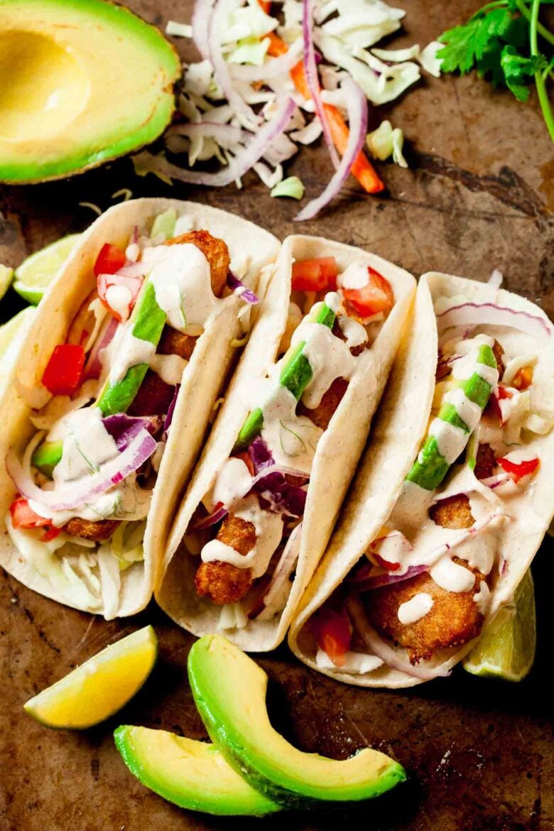 Three crispy Baja Fish Tacos in flour tortillas topped with shredded cabbage, sliced avocado, red onion, diced tomatoes, and creamy sauce.