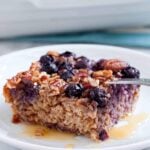 A square slice of gluten-free Baked Blueberry Oatmeal with pecans sits on a white plate, topped with a drizzle of syrup.