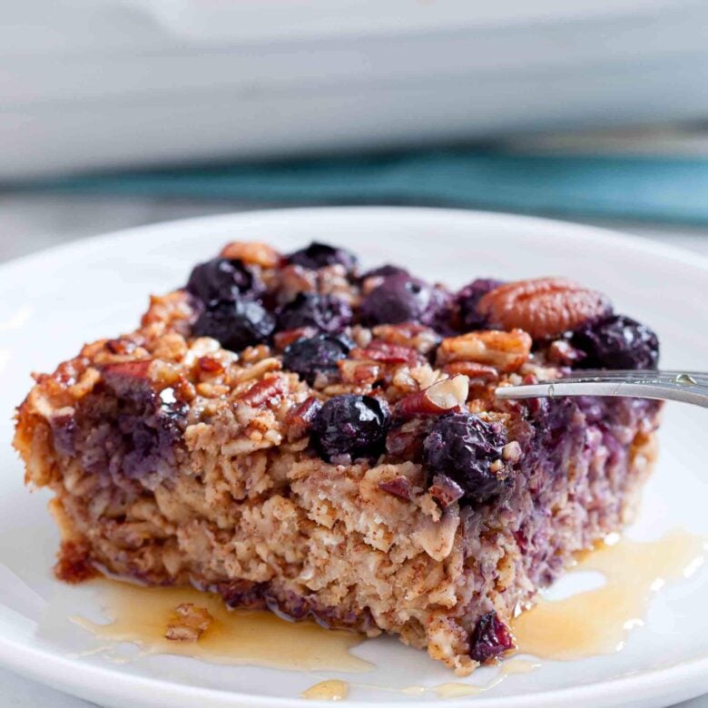 A square slice of gluten-free Baked Blueberry Oatmeal with pecans sits on a white plate, topped with a drizzle of syrup.