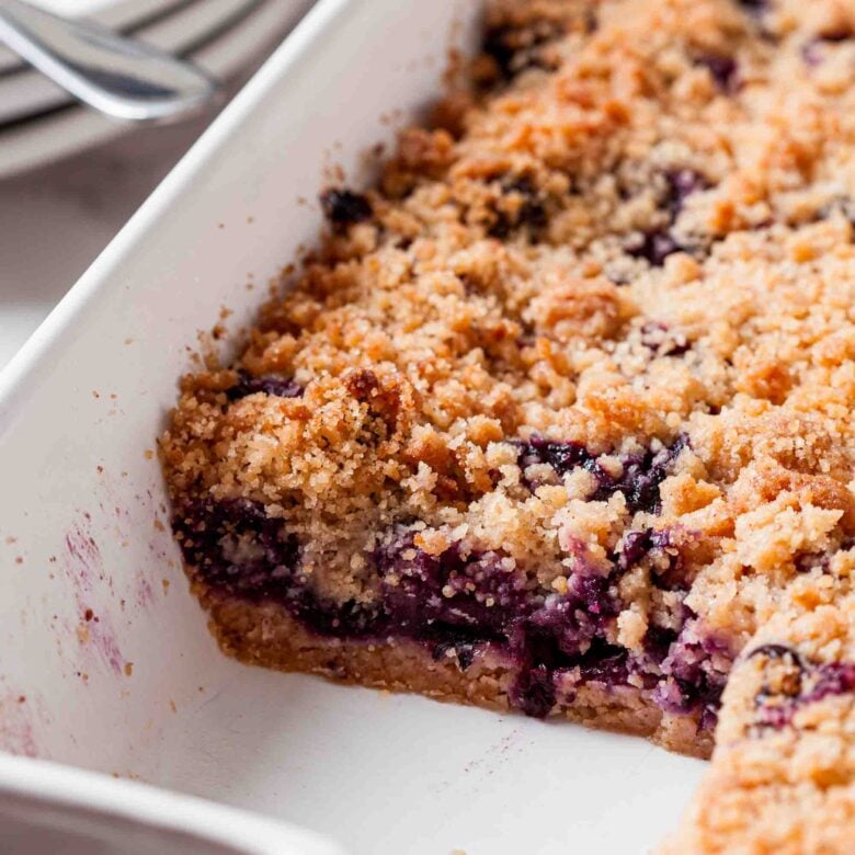 A close-up of a baked blueberry crumb bars dessert in a white rectangular dish, showing a missing corner slice.