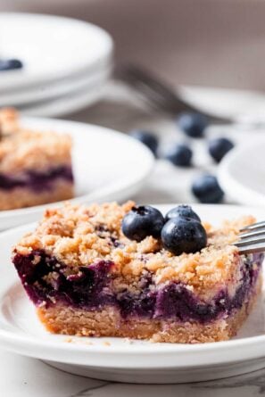 A square slice of blueberry crumb bar, reminiscent of blueberry pie dessert, is topped with two fresh blueberries on a white plate, a fork to the side and more blueberries in the background.