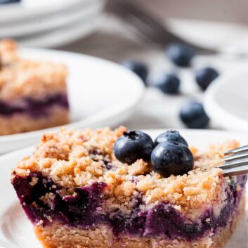 A square slice of blueberry crumb bar, reminiscent of blueberry pie dessert, is topped with two fresh blueberries on a white plate, a fork to the side and more blueberries in the background.