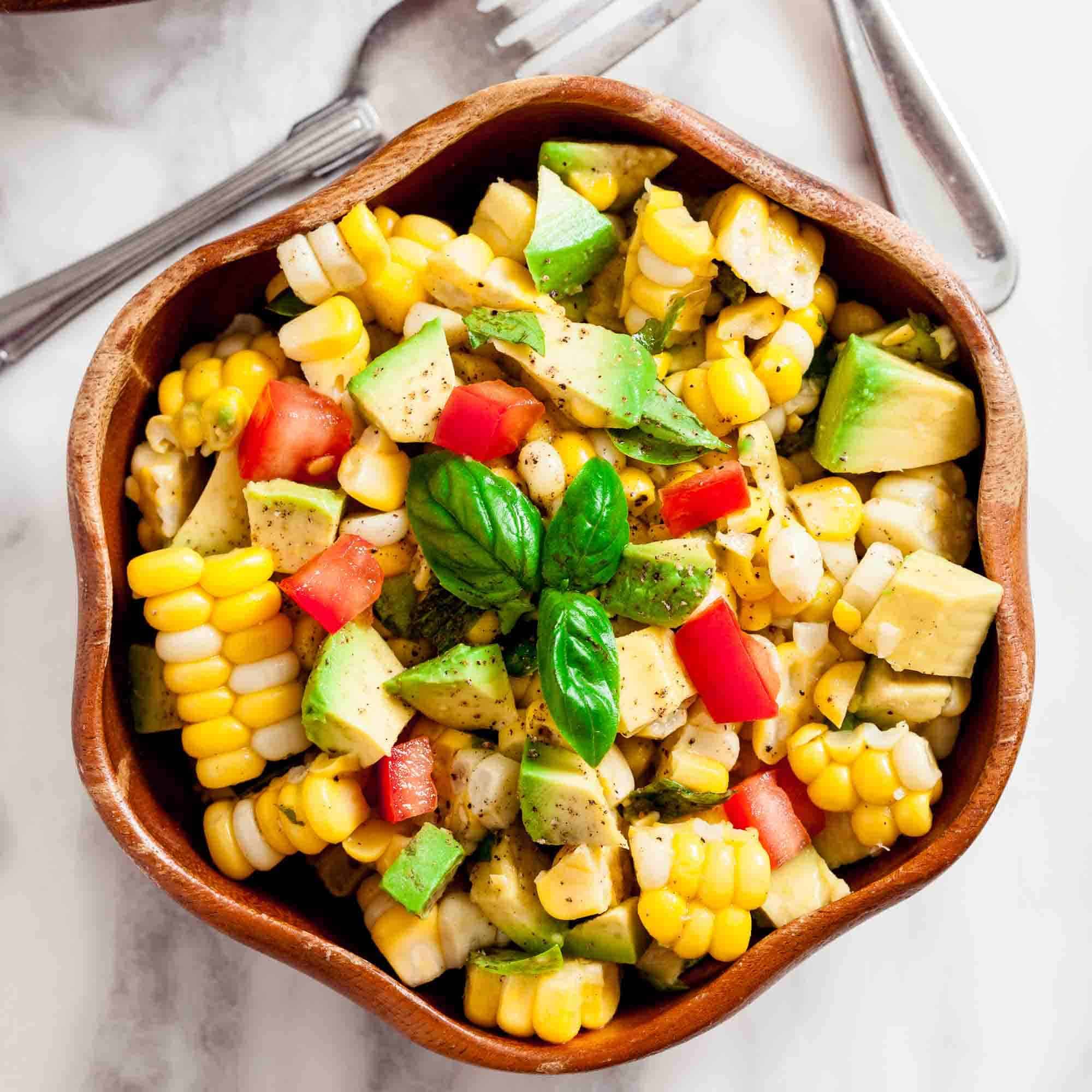 A wooden bowl filled with a healthy Corn Avocado Salad featuring corn, avocado, tomato, and fresh basil.