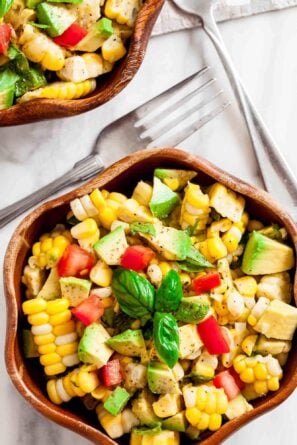 A wooden bowl filled with a healthy Corn Avocado Salad featuring tomato and fresh basil, with Tex-Mex flair, sits beside two forks on a white surface.