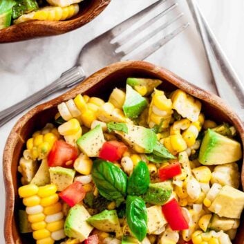 A wooden bowl filled with a healthy Corn Avocado Salad featuring tomato and fresh basil, with Tex-Mex flair, sits beside two forks on a white surface.