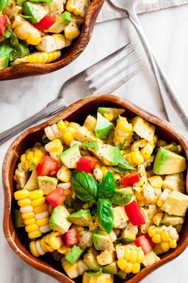 A wooden bowl filled with a healthy Corn Avocado Salad featuring tomato and fresh basil, with Tex-Mex flair, sits beside two forks on a white surface.