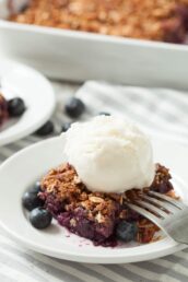 A serving of healthy blueberry crumble topped with a scoop of vanilla ice cream, garnished with fresh blueberries on a white plate with a fork.
