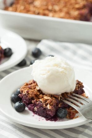 A serving of healthy blueberry crumble topped with a scoop of vanilla ice cream, garnished with fresh blueberries on a white plate with a fork.