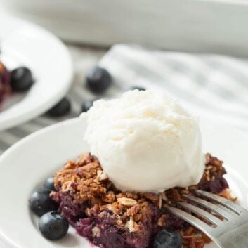 A serving of healthy blueberry crumble topped with a scoop of vanilla ice cream, garnished with fresh blueberries on a white plate with a fork.