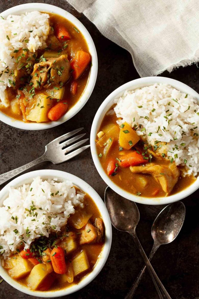 Three bowls of Japanese chicken curry over rice, featuring chunks of potato, carrot, and meat, are placed on a dark surface with two spoons, a fork, and a napkin nearby.