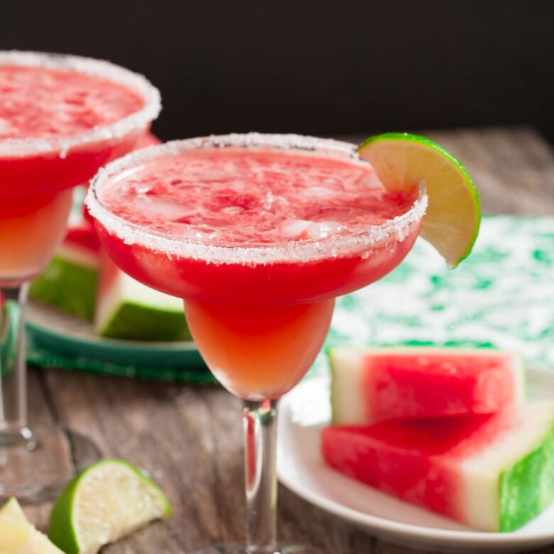 A salt-rimmed margarita glass filled with a refreshing red Watermelon Margarita, garnished with a lime wedge. Slices of watermelon and lime are visible in the background.