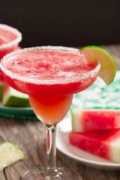 A salt-rimmed margarita glass filled with a refreshing red Watermelon Margarita, garnished with a lime wedge. Slices of watermelon and lime are visible in the background.
