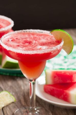 A salt-rimmed margarita glass filled with a refreshing red Watermelon Margarita, garnished with a lime wedge. Slices of watermelon and lime are visible in the background.
