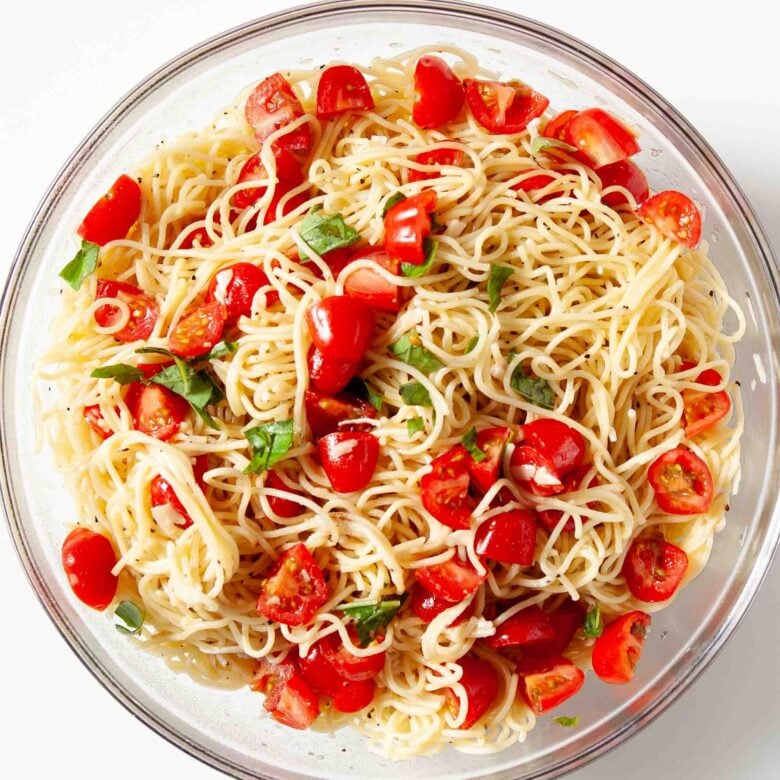 Overhead shot of glass bowl with bruschetta chicken pasta ingredients being tossed together.