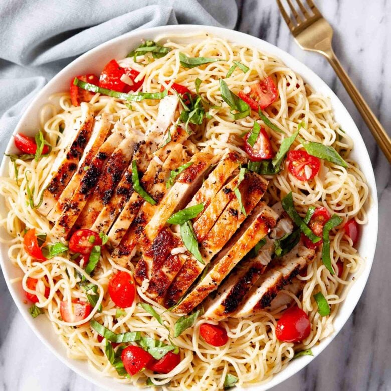 Overhead shot of a white bowl of bruschetta chicken pasta with angel hair, cherry tomatoes, and fresh basil sits on a marble surface.