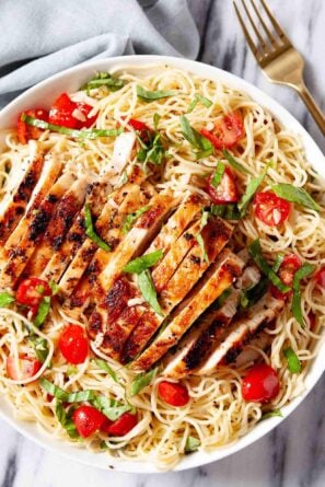 A white bowl of bruschetta chicken pasta with angel hair, cherry tomatoes, and fresh basil sits on a marble surface with a fork nearby.