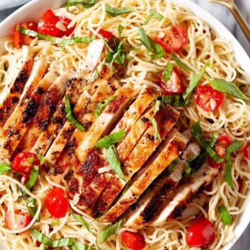 A white bowl of bruschetta chicken pasta with angel hair, cherry tomatoes, and fresh basil sits on a marble surface with a fork nearby.