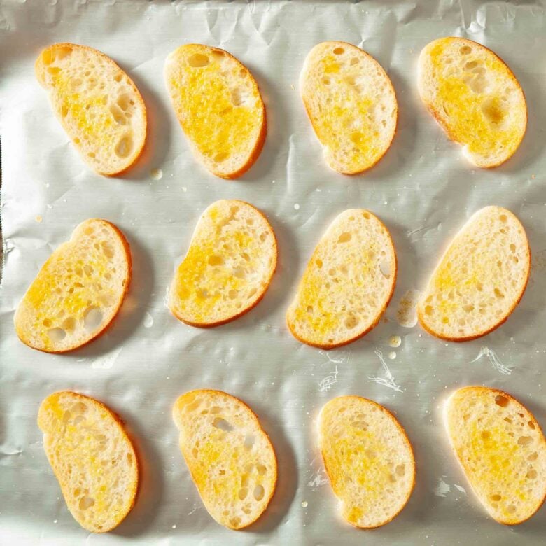 Garlic oil brushed over baguette slices on a baking sheet lined with foil.