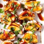 Sliced baguette topped with goat cheese, peach slices, chopped basil, and a drizzle of balsamic glaze arranged on a white platter to make peach crostini at home.